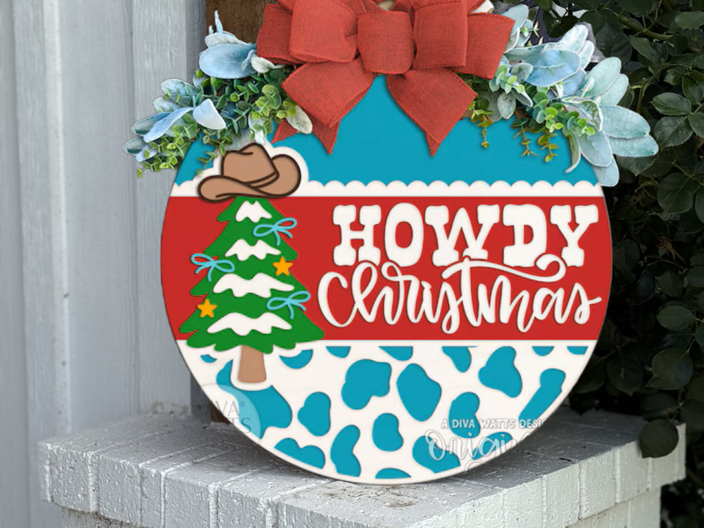 A round sign with blue cow spots, a Christmas tree with a cowboy hat, and the text Howdy Christmas in white and red, decorated with greenery, blue flowers, and a red bow, sits on a white brick ledge.