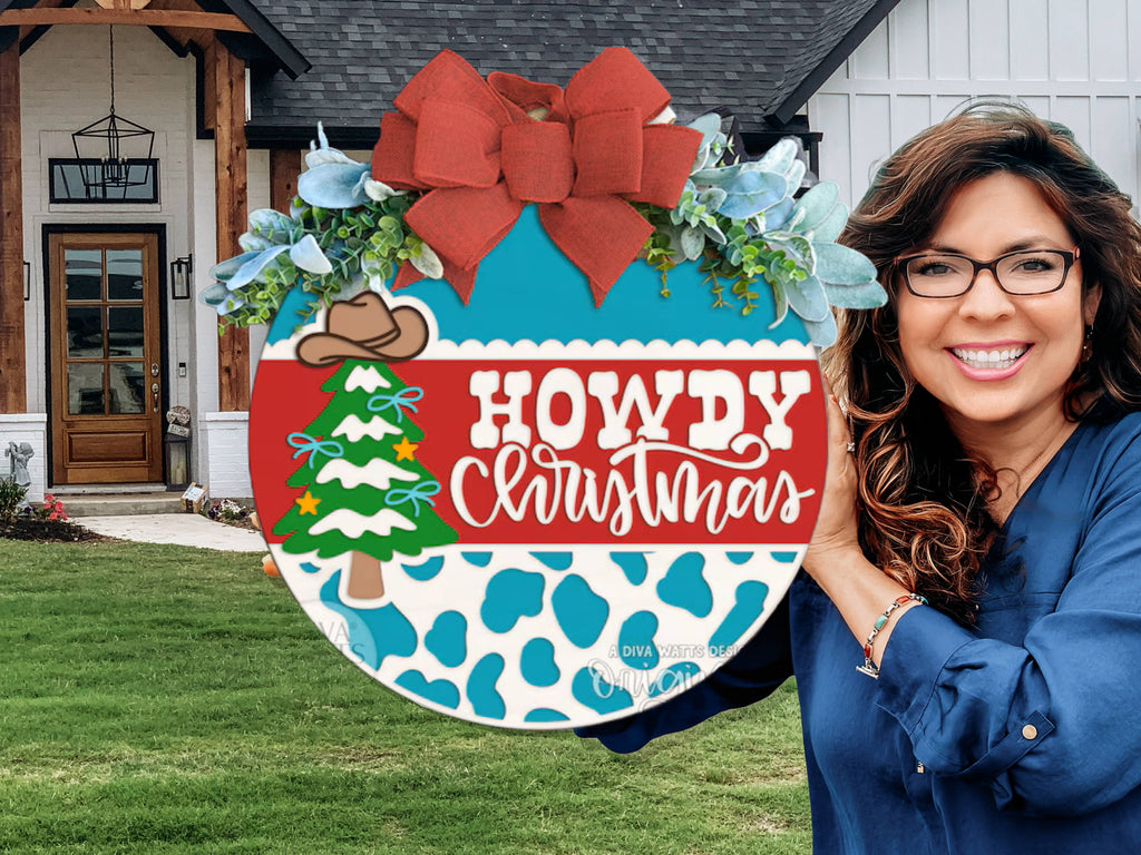 A smiling woman stands outside a house, holding a festive round sign with a red bow, greenery, a cowboy hat, a decorated tree, and the text “Howdy Christmas” above a blue cow print pattern.
