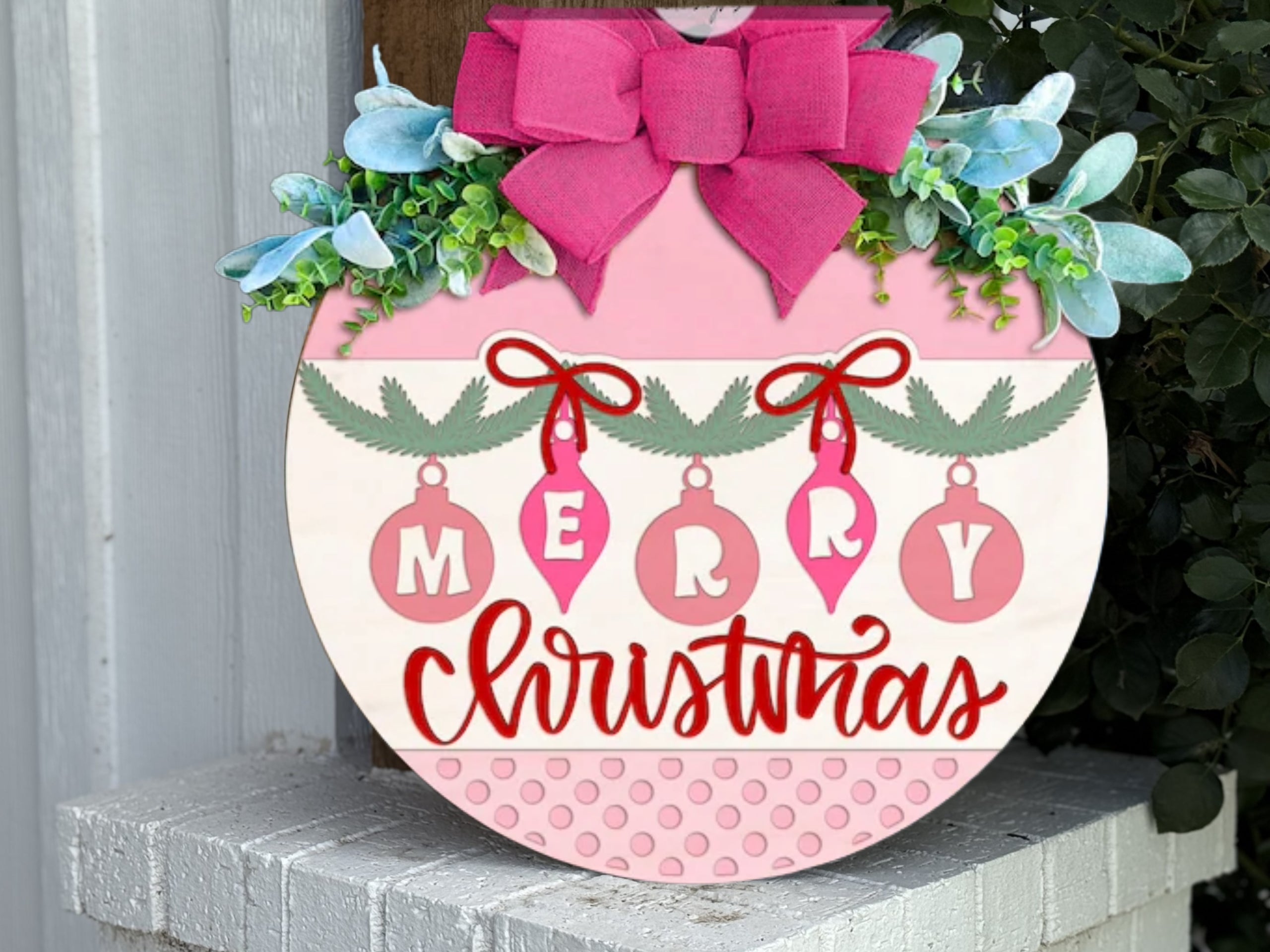 A round Christmas sign decorated with a large pink bow, greenery, and hanging ornaments spelling “MERRY,” with “Christmas” written in red script below. The sign rests on a white brick ledge.
