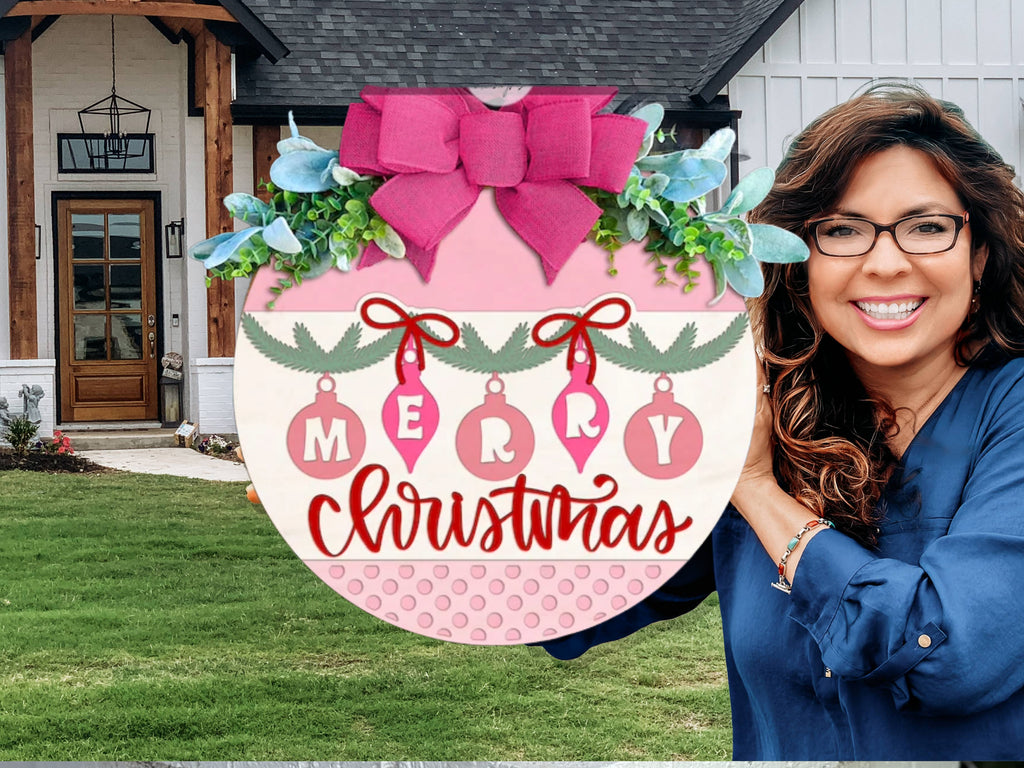 A smiling woman with wavy brown hair and glasses holds a large, round Merry Christmas sign decorated with a pink bow, ornaments, and greenery, standing outside in front of a modern house.