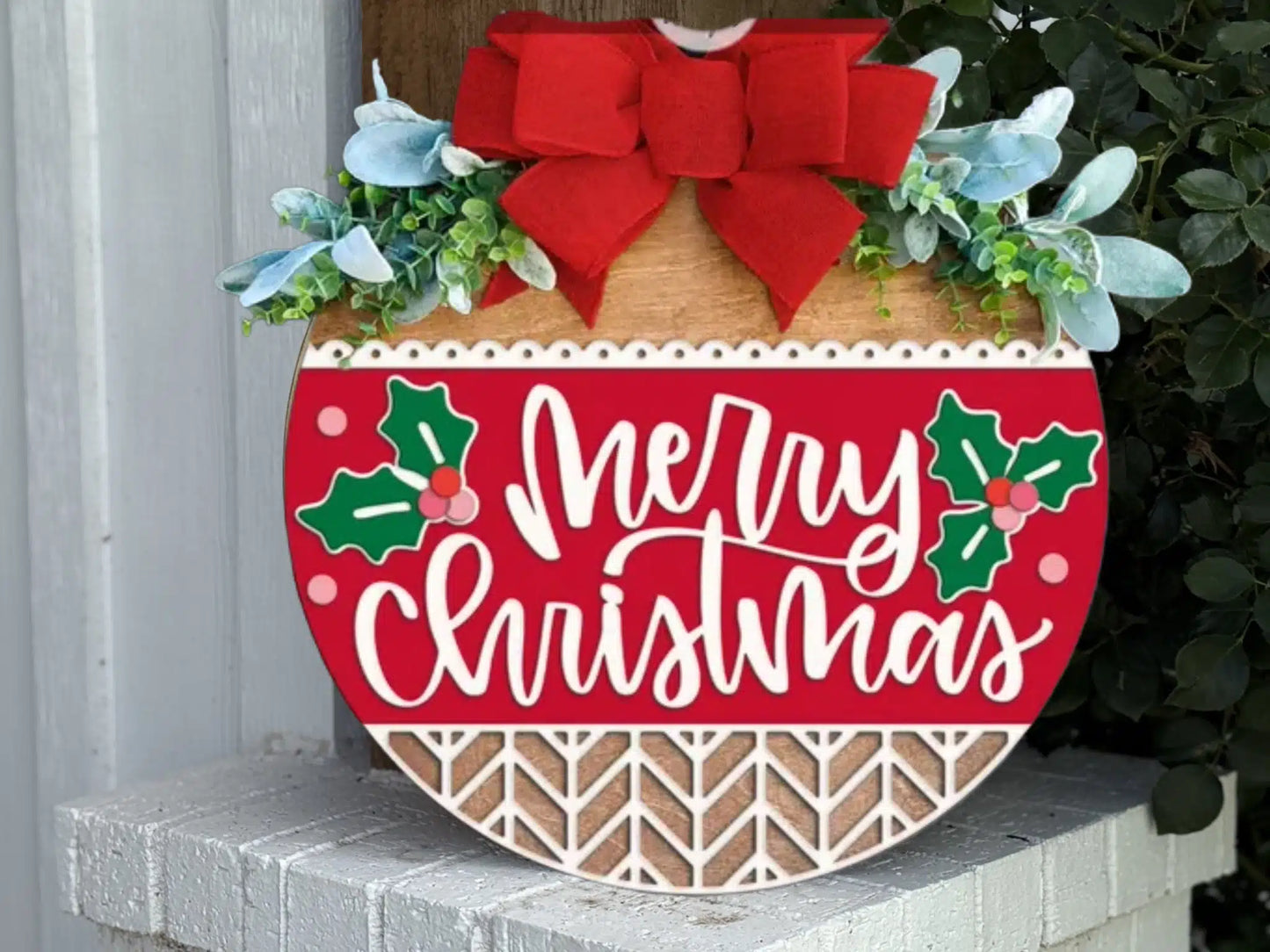A round wooden sign decorated with greenery, a red bow, and holly accents, featuring the words Merry Christmas in white script on a red background, leaning on a white brick ledge.
