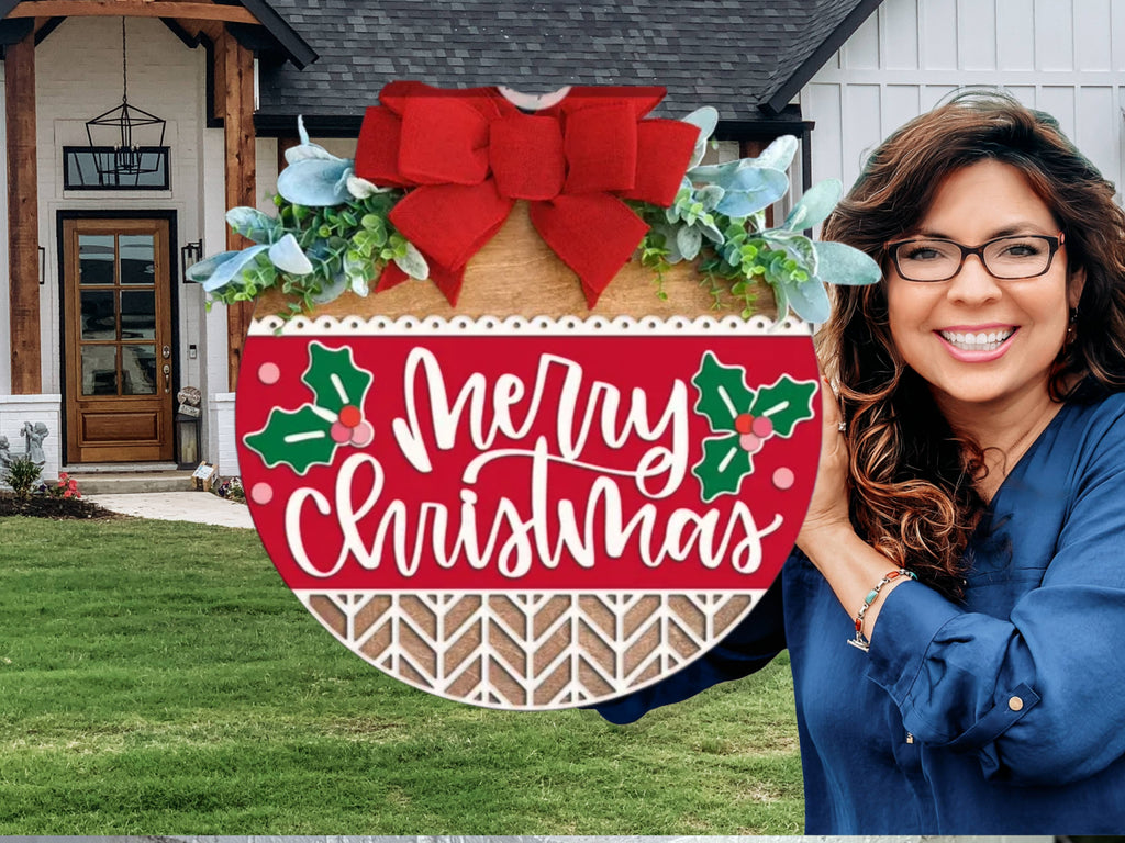 A smiling woman with long brown hair and glasses holds a round wooden sign decorated with a red bow and greenery. The sign reads Merry Christmas and features holly designs. A modern house with a lawn is in the background.