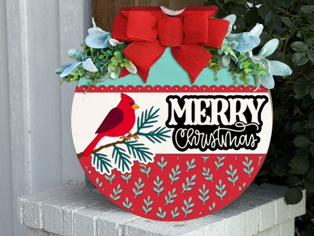 A round Christmas sign decorated with a red cardinal, greenery, and white flowers. It features a large red bow on top and the words Merry Christmas in bold letters across the center.