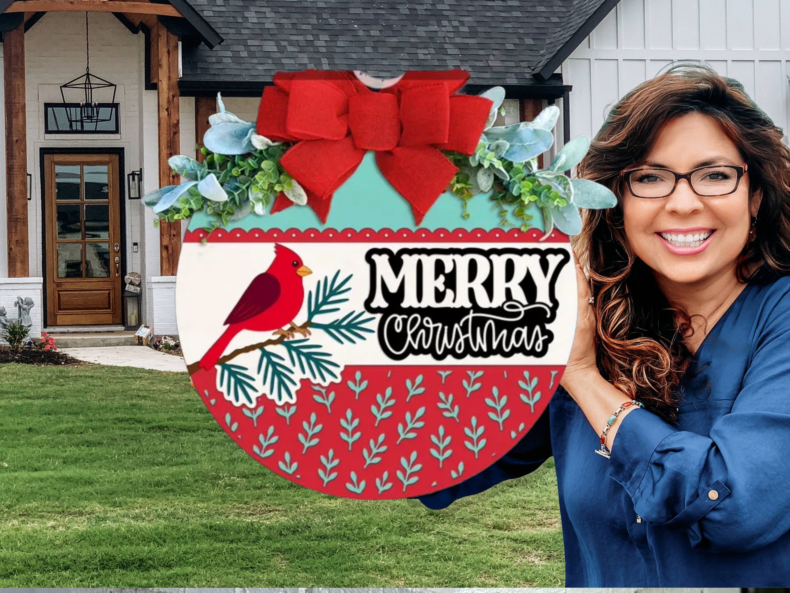 A smiling woman holding a round Christmas sign that reads “Merry Christmas,” decorated with a red bow, greenery, a red cardinal, and pine branches, standing outside in front of a house.