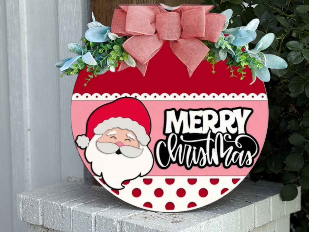 A round Christmas sign with a cartoon Santa, the words “Merry Christmas,” red and white polka dots, a pink bow, and green leaves sits on a white ledge beside greenery.
