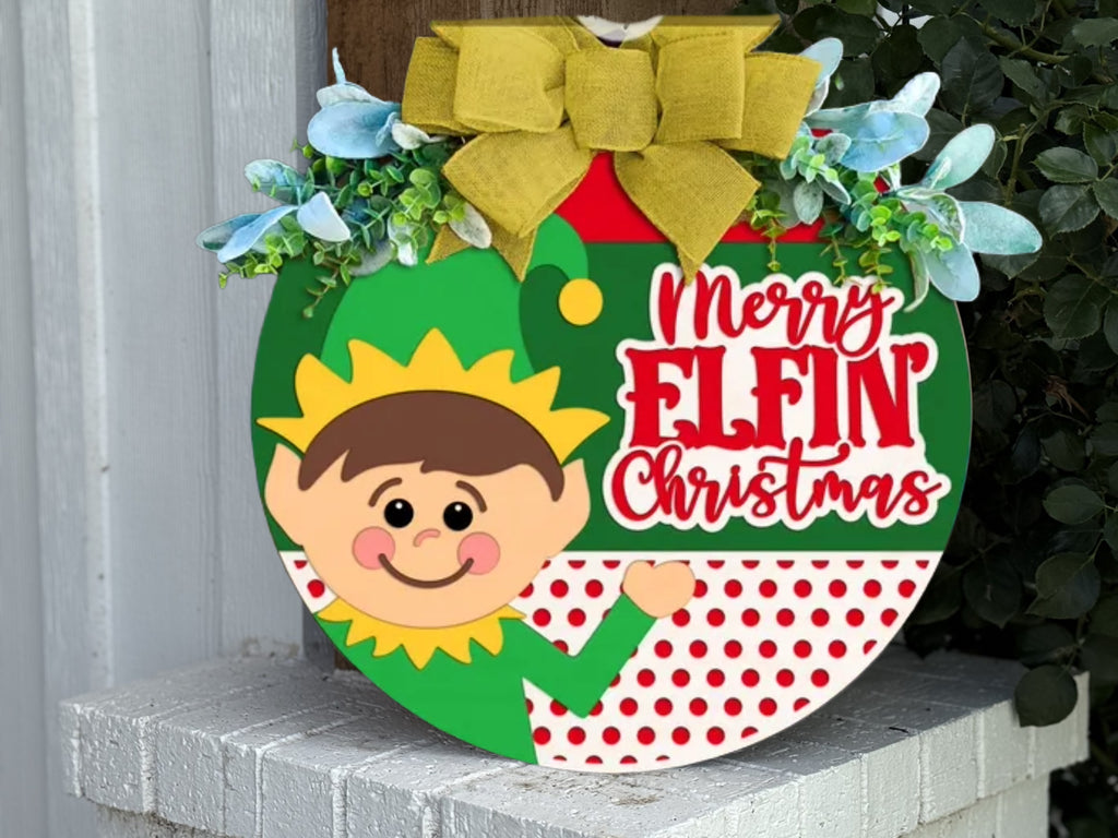 A round holiday sign decorated with a yellow bow, greenery, and flowers features a smiling cartoon elf and the words Merry Elfin Christmas in red letters on a red and white polka dot background.