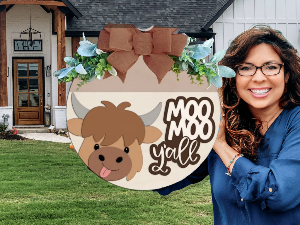 A smiling woman with wavy brown hair and glasses holds a circular sign with a cartoon cow, greenery, a large brown bow, and the words Moo Moo Y’all in front of a modern house.