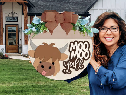 A smiling woman with wavy brown hair and glasses holds a circular sign with a cartoon cow, greenery, a large brown bow, and the words Moo Moo Y’all in front of a modern house.