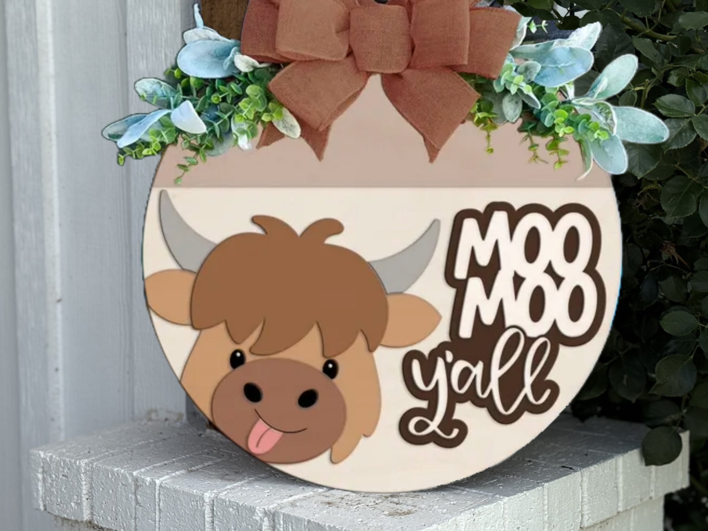A round wooden sign with a cartoon cow, greenery, a burlap bow on top, and the words “Moo Moo y’all.” The sign sits on a white brick ledge next to some green leaves.