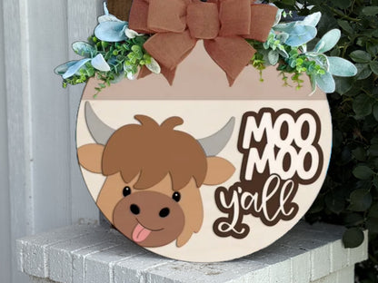 A round wooden sign with a cartoon cow, greenery, a burlap bow on top, and the words “Moo Moo y’all.” The sign sits on a white brick ledge next to some green leaves.
