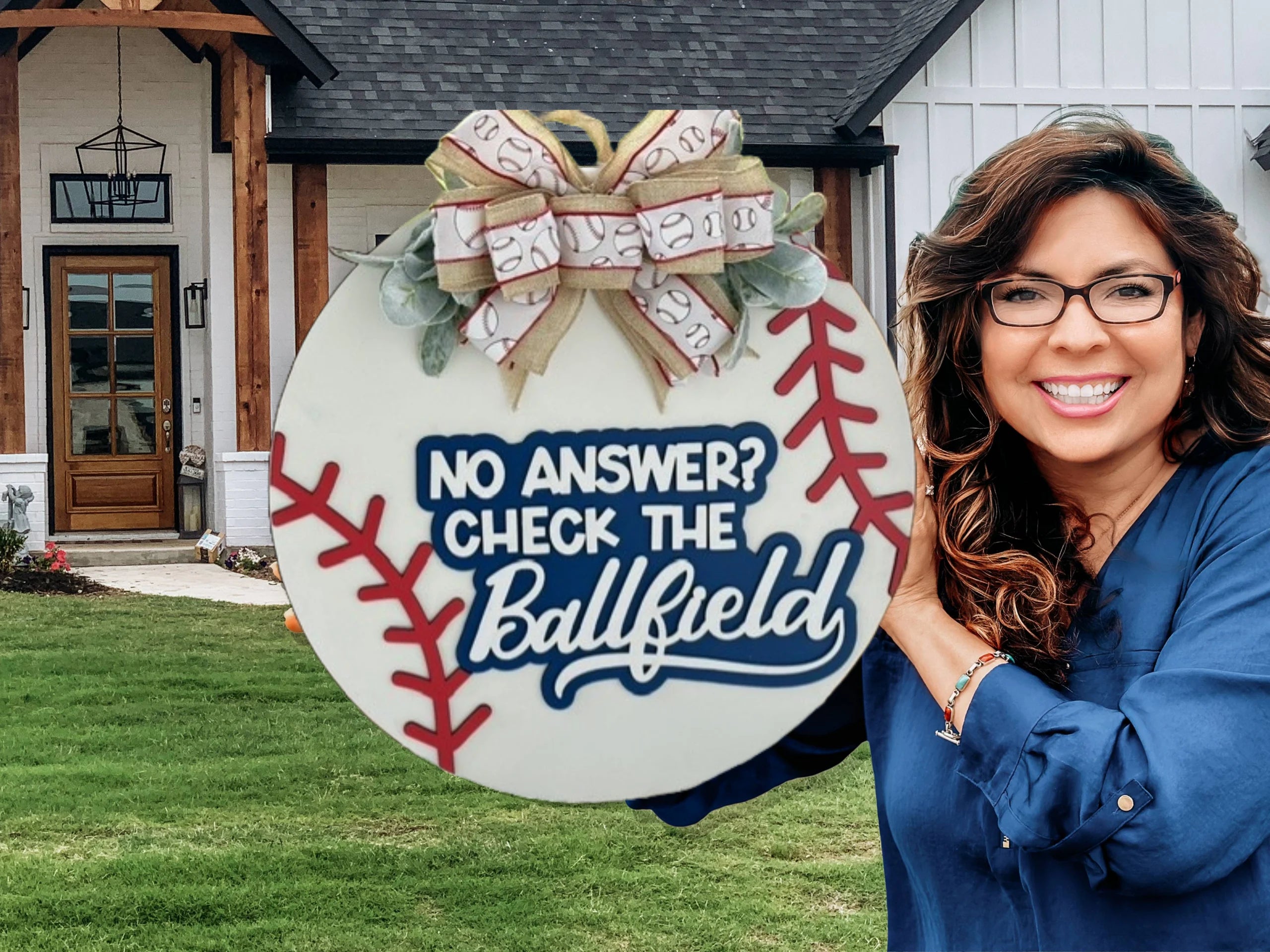 A woman in glasses and a blue shirt smiles while holding a round, baseball-themed sign with the text No answer? Check the Ballfield. The sign is decorated with a bow and greenery. In the background, theres a house with a wooden door, black roof, and manicured lawn.