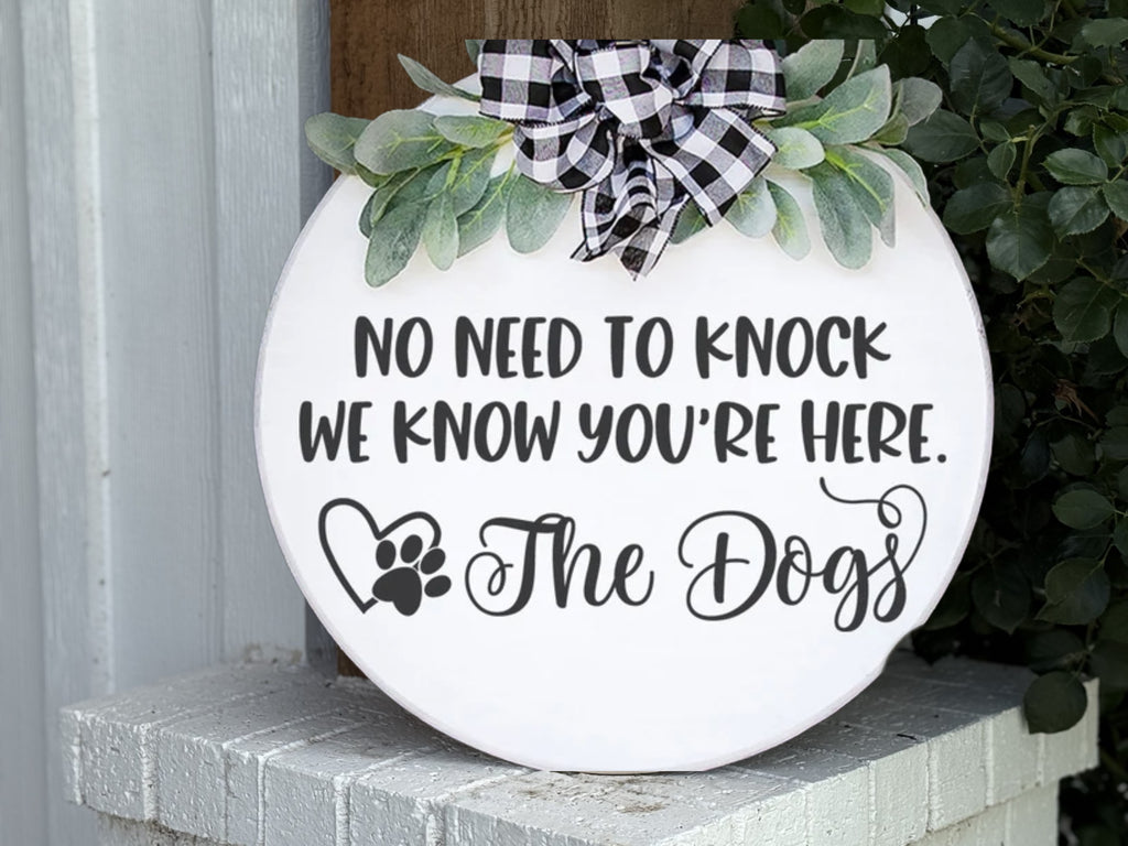 A round white sign with greenery and a black-and-white plaid bow reads, “No need to knock, we know you’re here. The Dogs,” with a heart and paw print graphic. The sign is propped on a white brick surface.