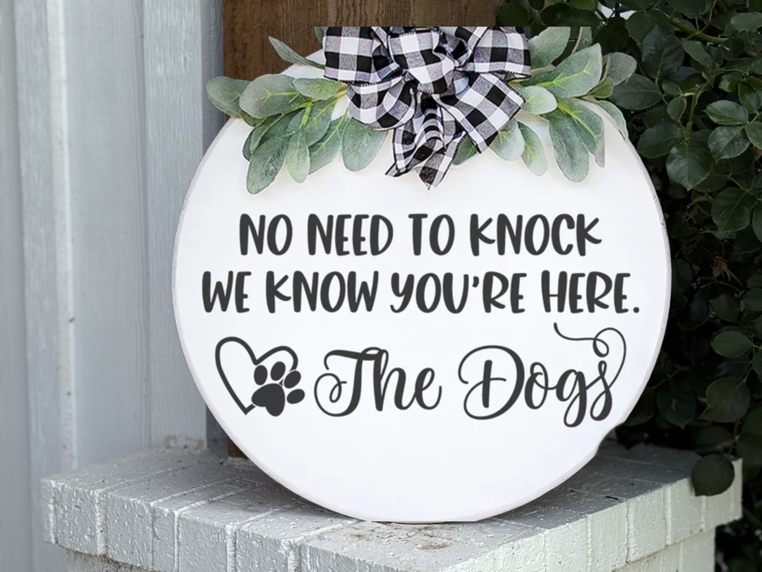 A round white sign with greenery and a black-and-white plaid bow reads, “No need to knock, we know you’re here. The Dogs,” with a heart and paw print graphic. The sign is propped on a white brick surface.