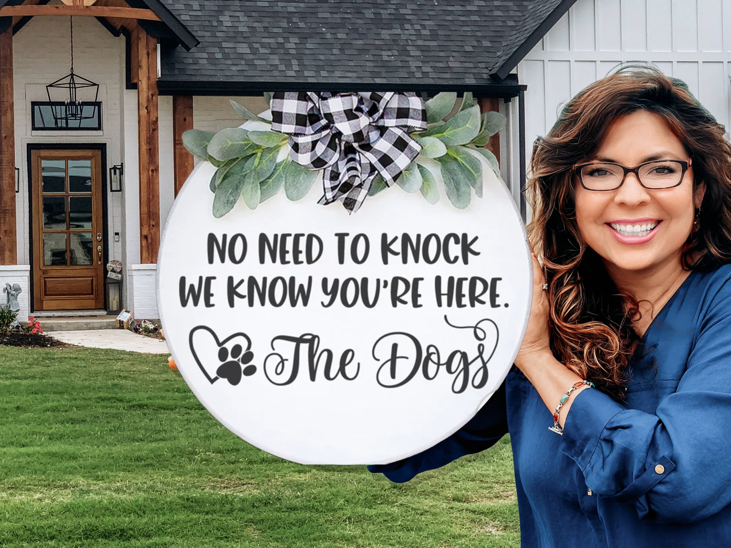 A smiling woman stands outside a house, holding a round sign decorated with greenery and a plaid ribbon. The sign reads, No need to knock, we know you’re here. The Dogs, with a heart and paw print.