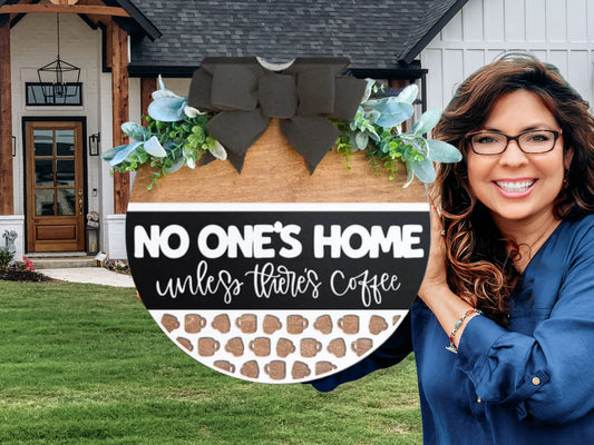 A smiling woman holds a decorative round sign in front of a house. The sign reads, NO ONES HOME unless theres coffee and is adorned with a black bow and greenery at the top.