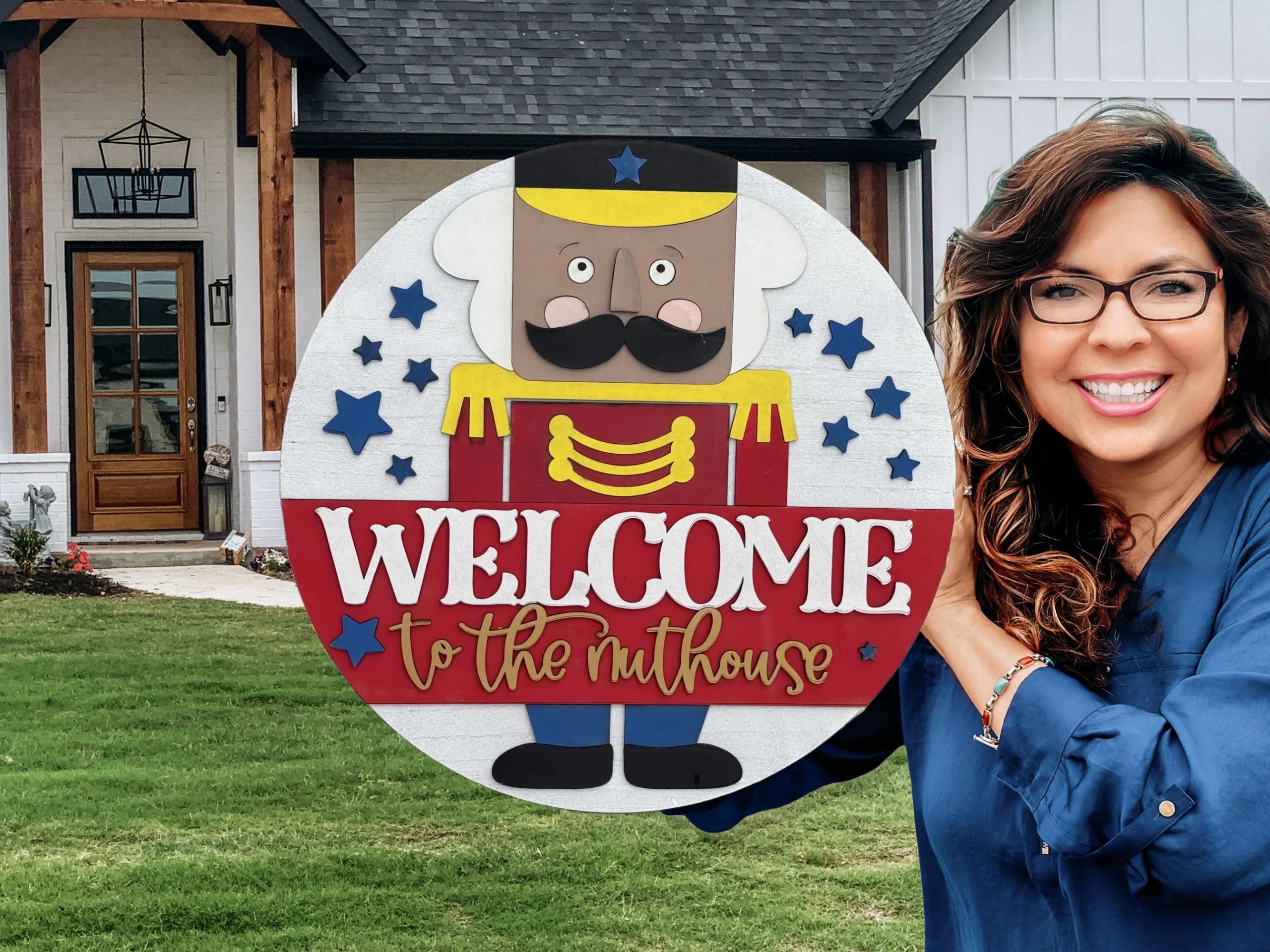 A smiling woman holds a round sign featuring a cartoon nutcracker and the text “WELCOME to the nuthouse” in front of a modern house with a lawn.