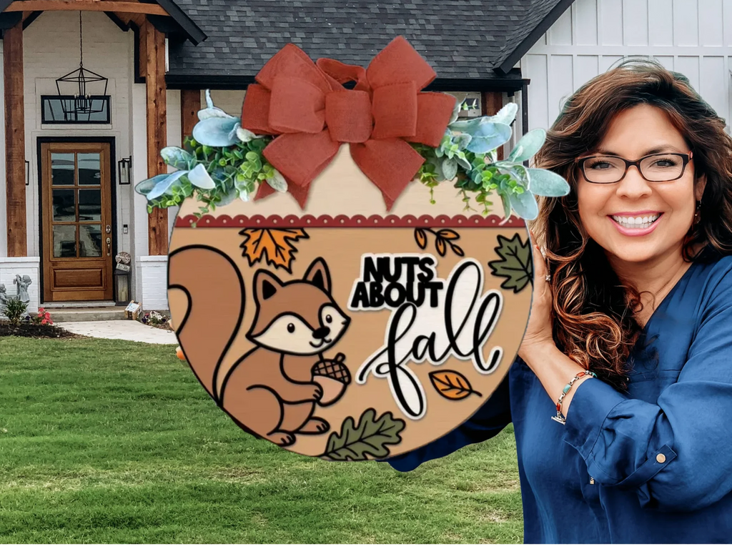 A smiling woman holds a round fall-themed sign with a squirrel, leaves, and the words Nuts About Fall in front of a modern house with white siding and wood accents.