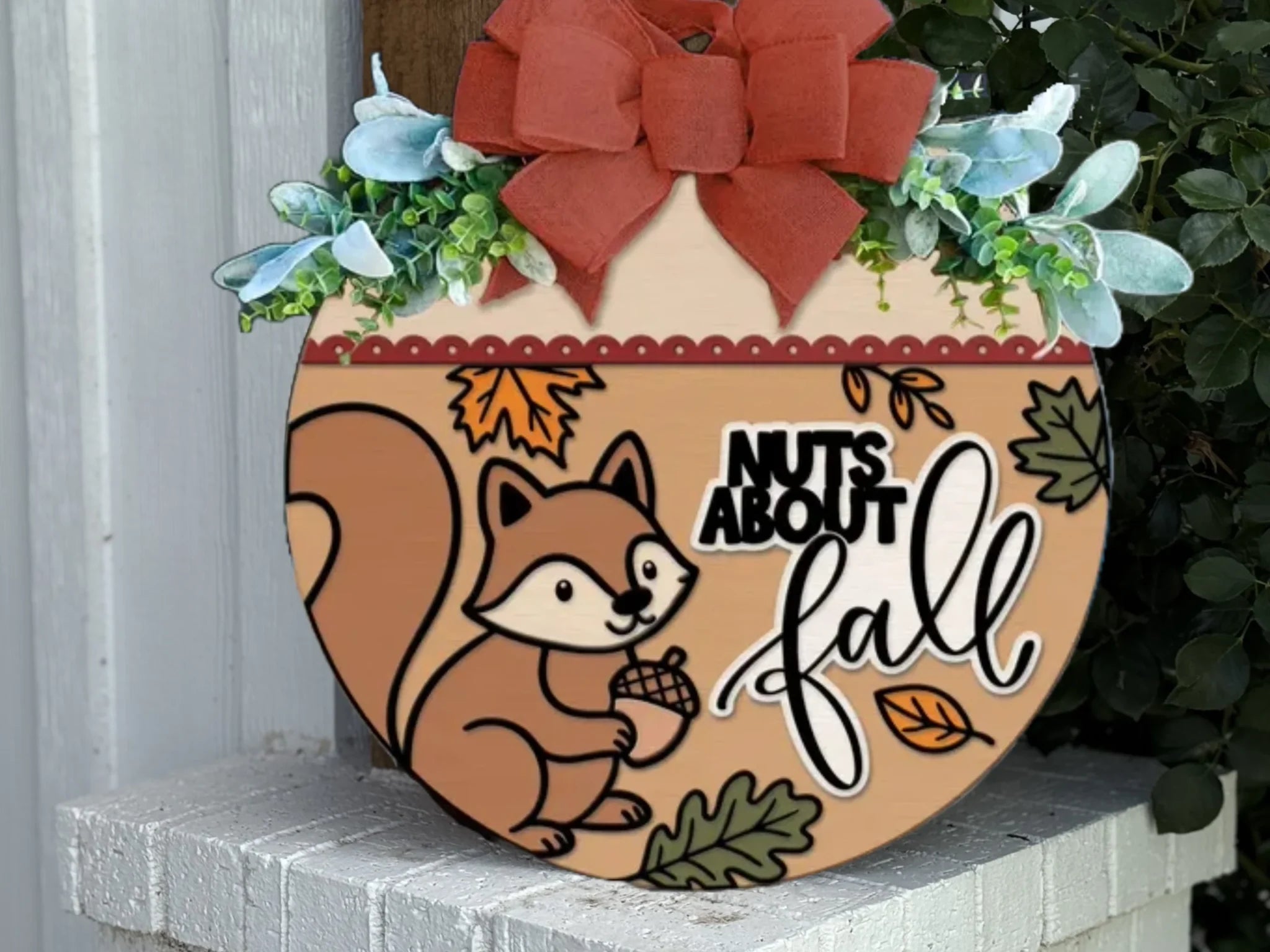 A round autumn-themed sign with a cartoon squirrel holding an acorn, surrounded by leaves. The sign reads Nuts about fall and is topped with a red bow and greenery. It rests on a white brick ledge.