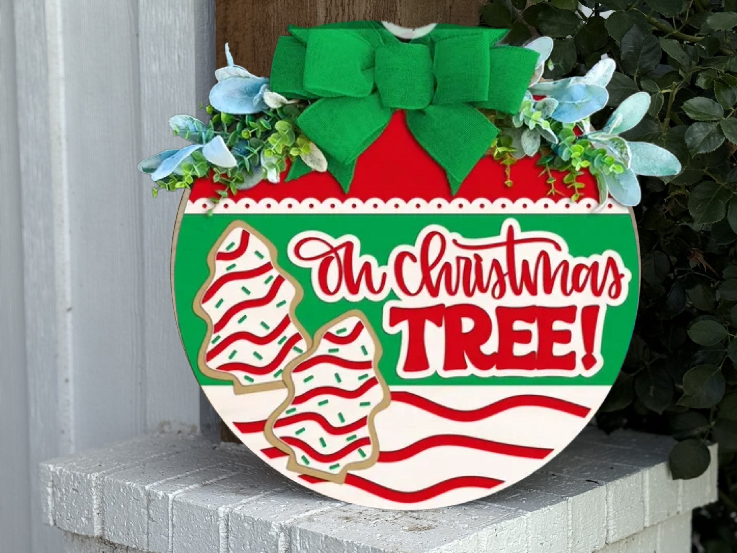 A round wooden sign decorated with a green bow, faux greenery, and two Christmas tree cookies, featuring the words Oh Christmas Tree! in festive red, green, and white colors. The sign is displayed outdoors.