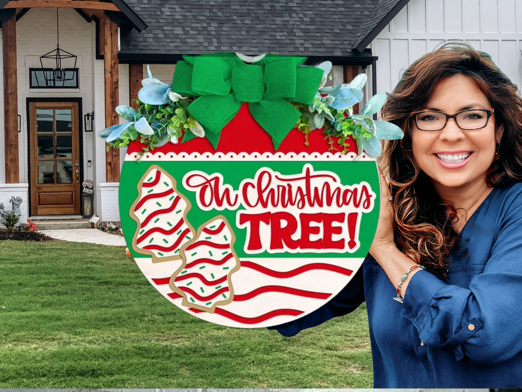 A smiling woman with wavy brown hair and glasses holds a festive Oh Christmas Tree! sign decorated with ornaments and greenery, standing in front of a modern house with a neatly trimmed lawn.