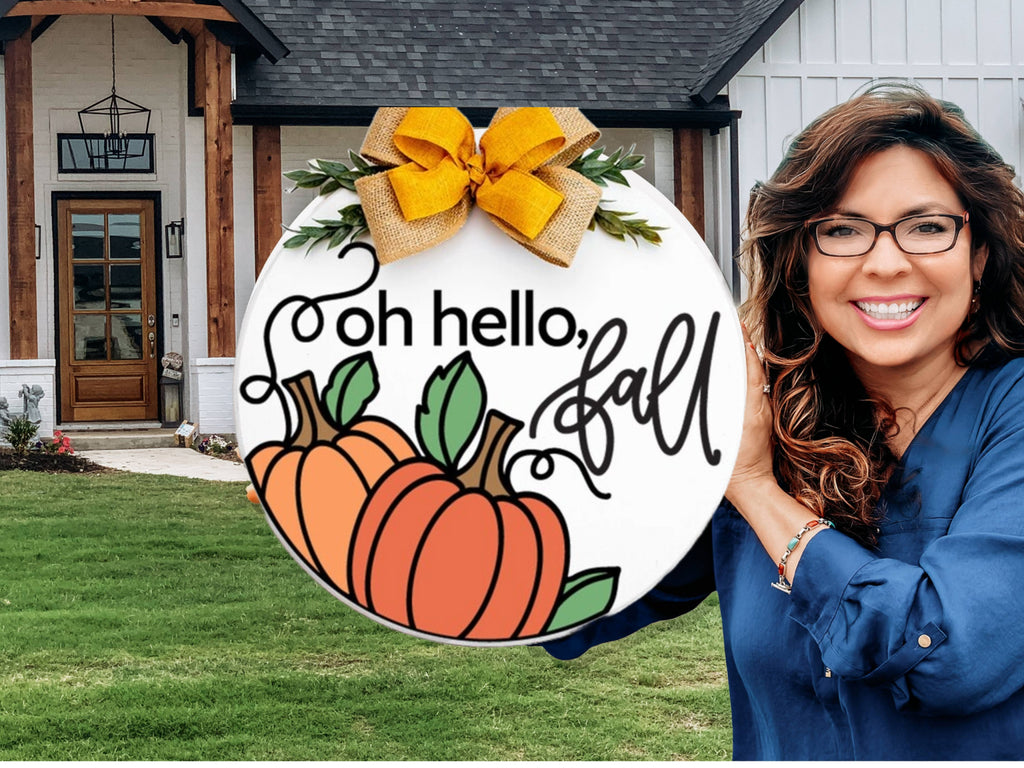 A smiling woman with wavy brown hair and glasses holds a round sign with pumpkins, leaves, a yellow bow, and the text oh hello, fall in front of a modern house with a green lawn.