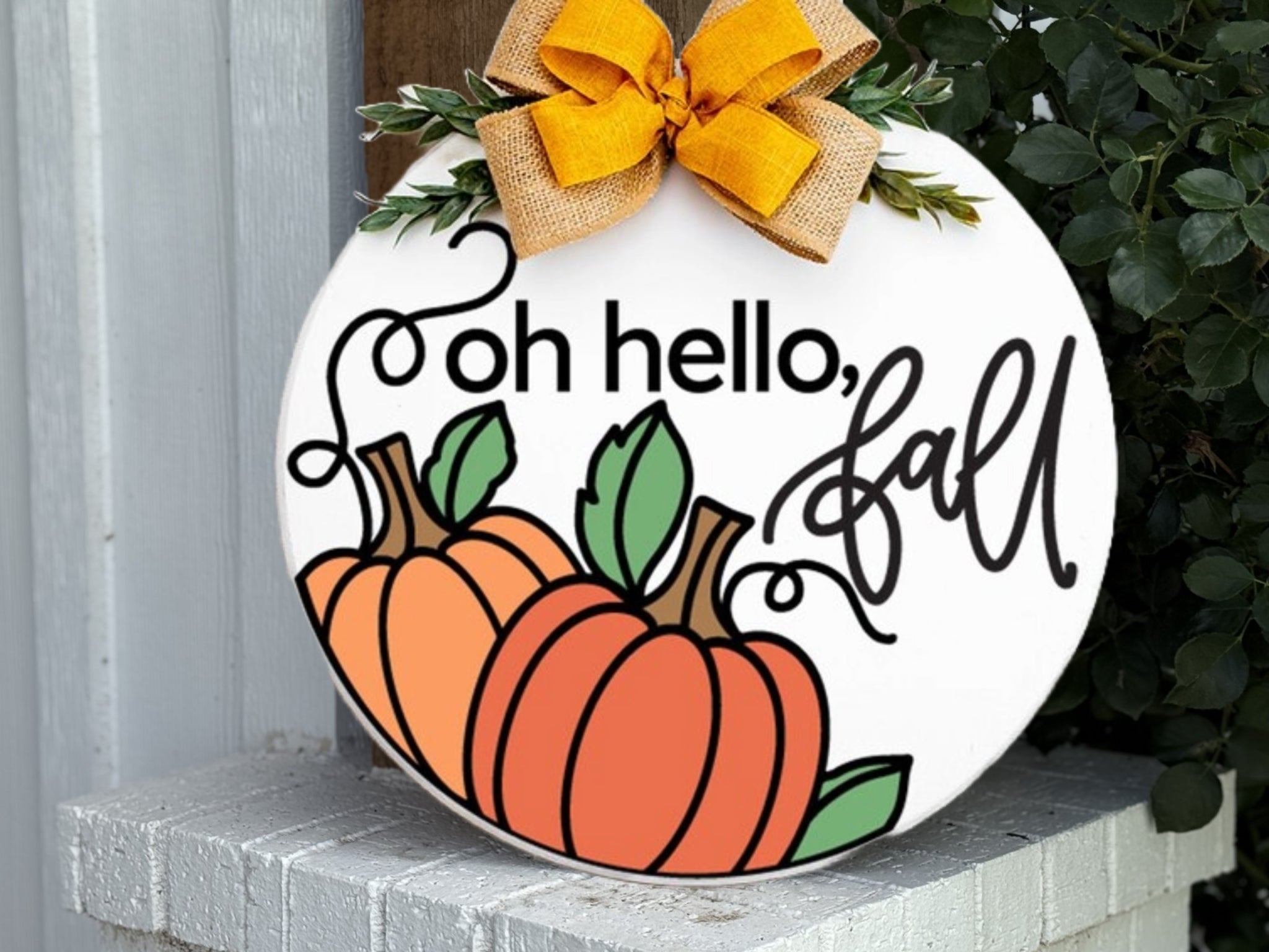 A round white sign decorated with two orange pumpkins, green leaves, and the words “oh hello, fall” in black script. A burlap and yellow ribbon bow is attached at the top. The sign rests on a white brick surface.