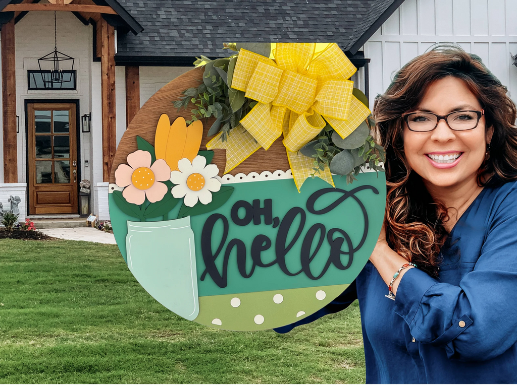 A smiling woman in glasses and a blue shirt holds a round wooden sign with colorful flowers, greenery, a big yellow plaid bow, and the words “oh, hello” in script. She stands on grass in front of a modern house with white siding and wood accents.