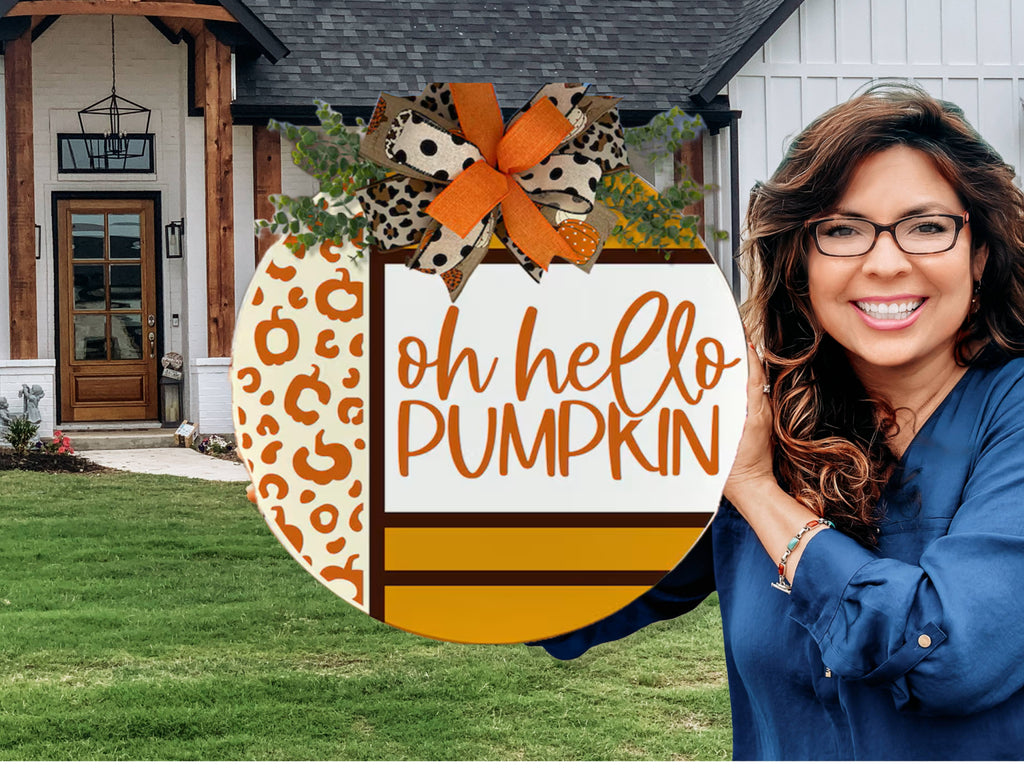 A smiling woman with long brown hair and glasses holds a round autumn-themed sign that reads oh hello PUMPKIN in front of a house with a wooden door and green lawn. The sign features orange, leopard print, and a decorative bow.