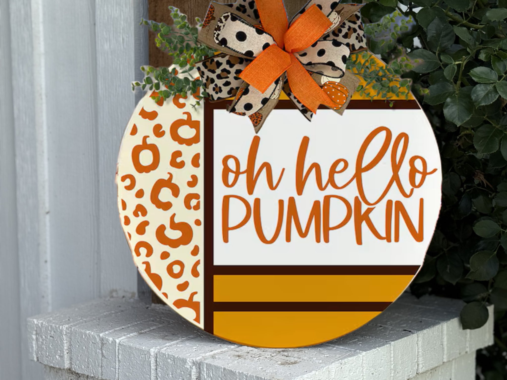 A round decorative sign with a cheetah print and stripes, featuring the text oh hello PUMPKIN in orange script. It is topped with a bow made of orange, burlap, and cheetah-print ribbons. The sign rests on a white brick.