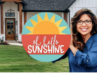 A smiling woman with long brown hair and glasses stands in front of a house, holding a round sign with a sun graphic and the words oh hello SUNSHINE written on it.
