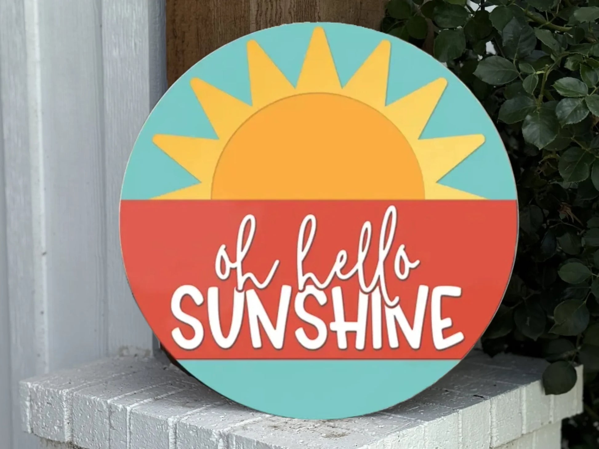A round sign with a sun rising over a red horizon, light blue sky, and the words oh hello SUNSHINE written in playful white letters. The sign rests on a white brick surface next to green leaves.