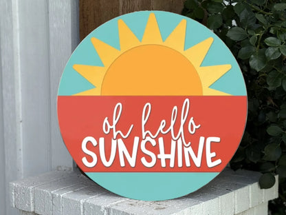A round sign with a sun rising over a red horizon, light blue sky, and the words oh hello SUNSHINE written in playful white letters. The sign rests on a white brick surface next to green leaves.