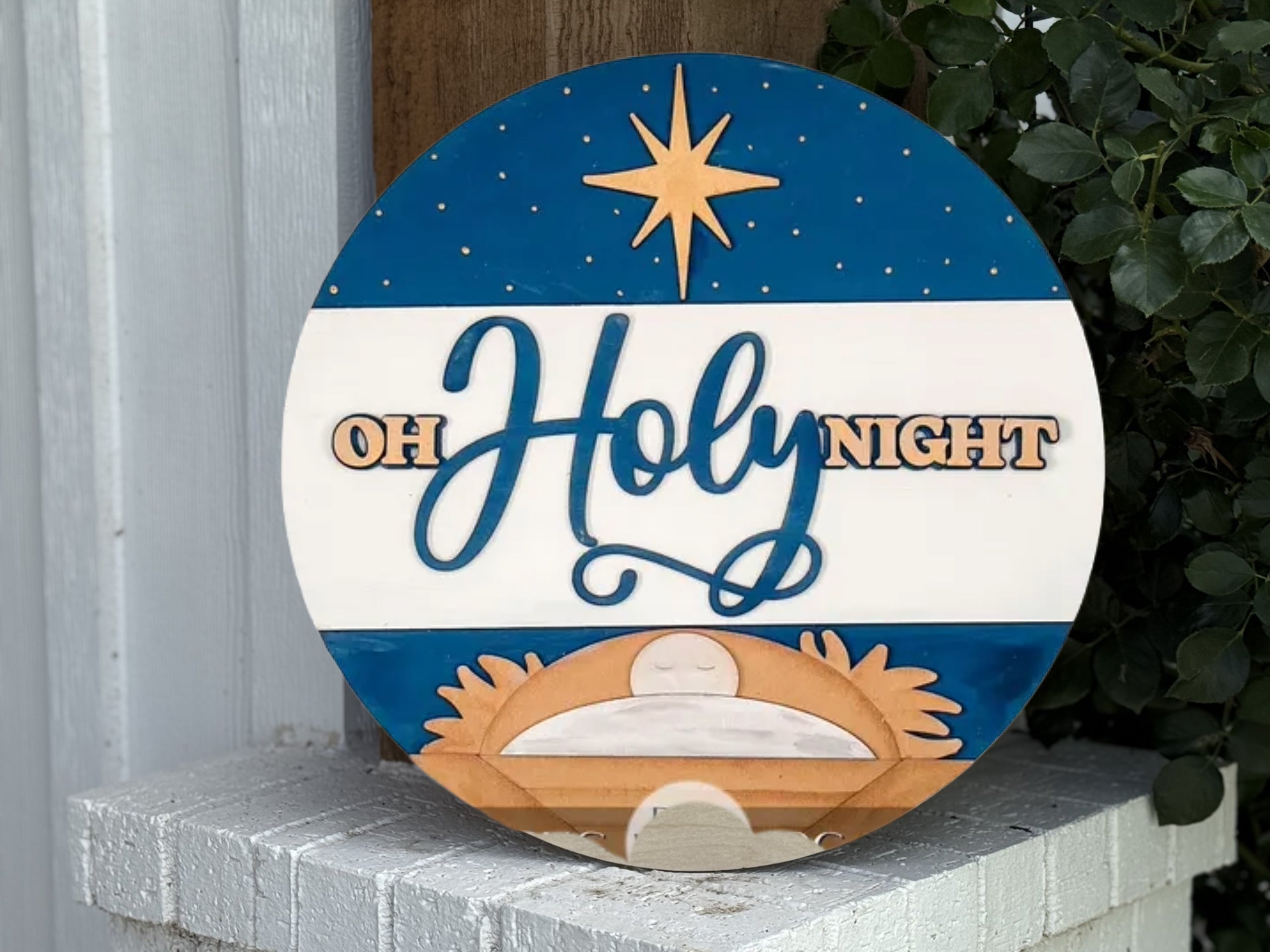 A round wooden sign with a nativity scene, a bright star in the night sky, and the words “OH HOLY NIGHT” in blue and gold letters. The sign is displayed outdoors next to greenery and a white wall.