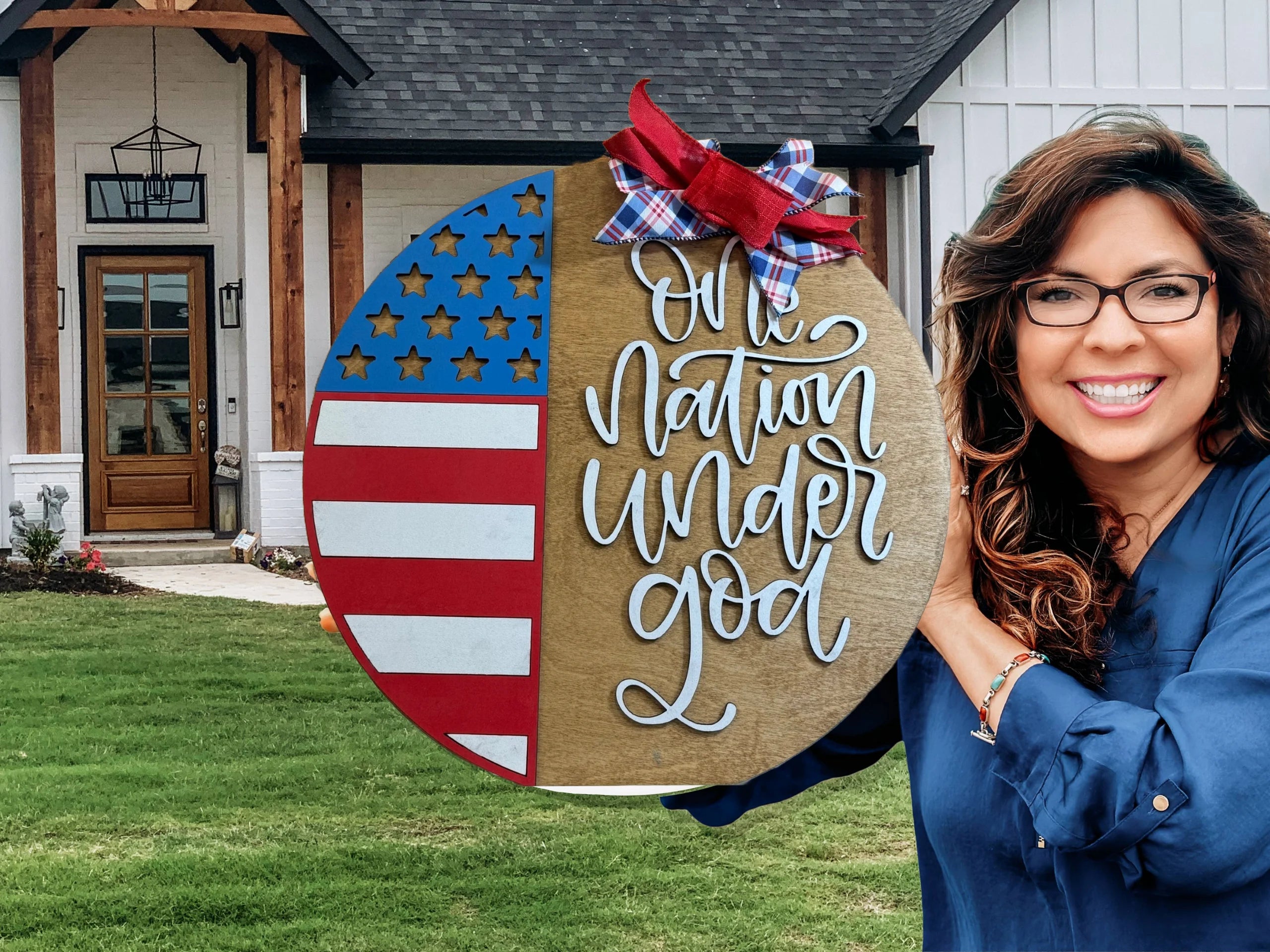 A smiling woman with glasses holds a decorative round sign featuring a U.S. flag design, a red plaid bow, and the words One nation under God, standing in front of a house with a lawn.