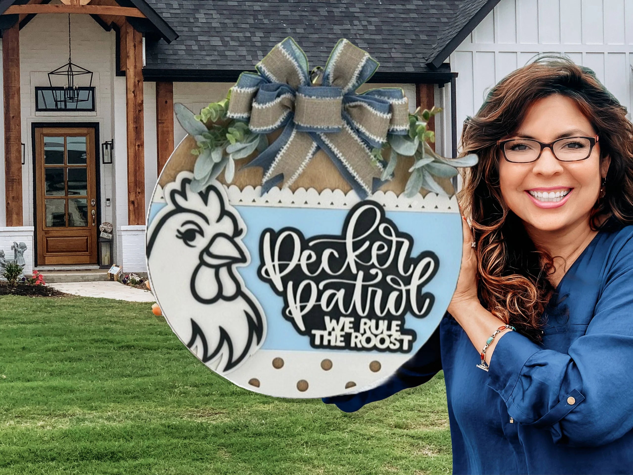 A smiling woman with glasses holds a decorative round sign featuring a chicken illustration and the words Pecker Patrol, We Rule The Roost, standing in front of a modern house with a wooden front door.