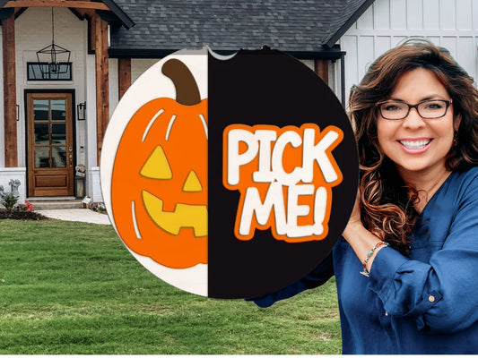 A smiling woman with long brown hair and glasses stands in front of a house, holding a round sign with a pumpkin illustration on one side and the words “PICK ME!” in bold letters on the other.