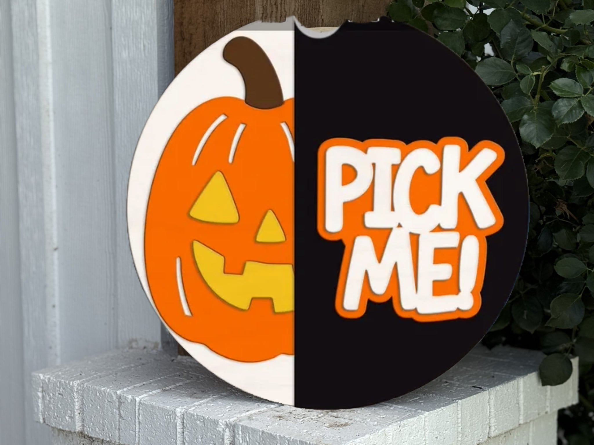 A round sign is divided in half; the left side shows a smiling cartoon jack-o-lantern, and the right side has the words “PICK ME!” in bold orange and white letters. The sign sits on a white brick ledge.