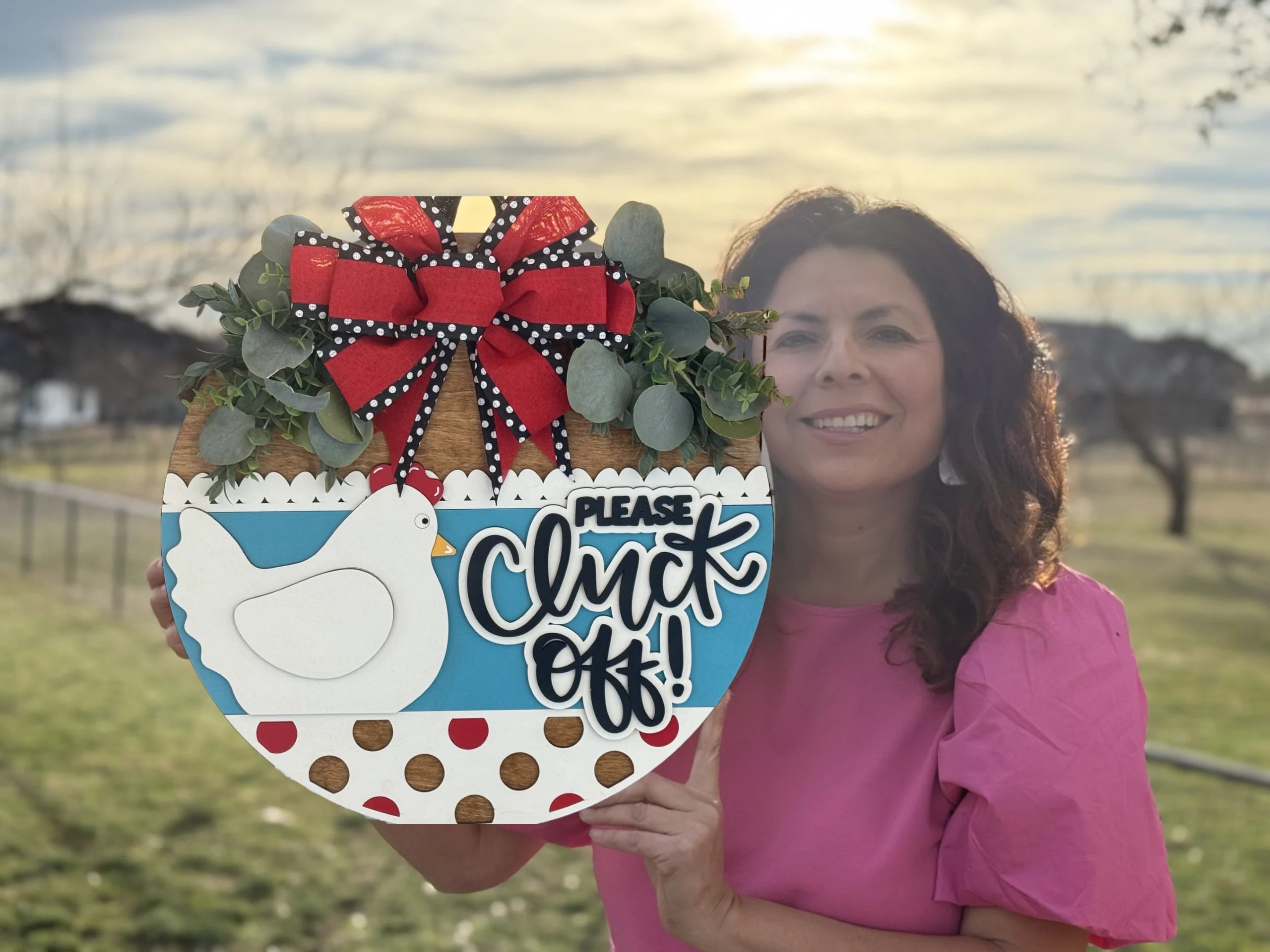A woman in a pink shirt holds a decorative round sign outdoors. The sign features a white hen, red and white polka dots, greenery, and a large red bow. It reads, Please Cluck Off! The sun is setting, casting a warm glow, with a blurred background of trees and fence.