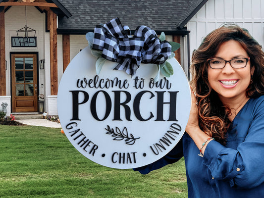 A smiling woman with glasses holds a round sign that reads Welcome to our porch. Gather. Chat. Unwind. The sign is decorated with a large black and white plaid bow. A house and green lawn are visible in the background.