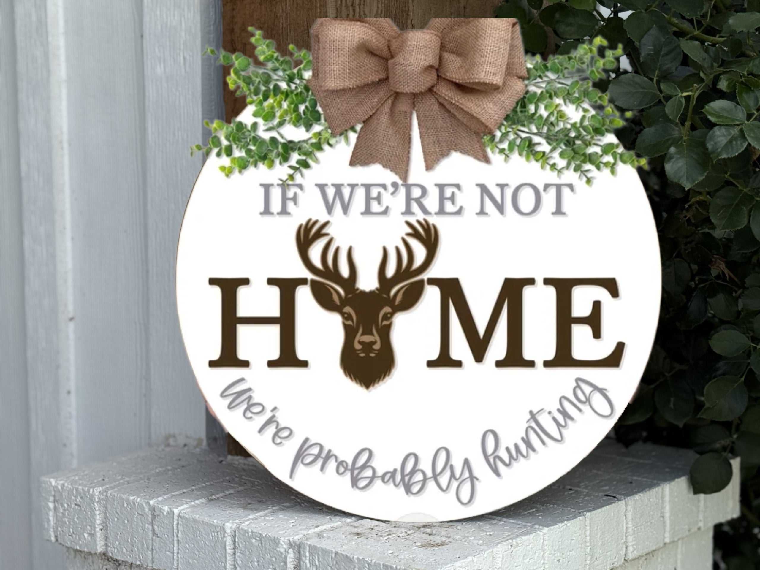 A round sign with a burlap bow and greenery reads, If we’re not HOME we’re probably hunting, with a deer head forming the “O” in “HOME.” The sign rests on a white brick surface near plants.