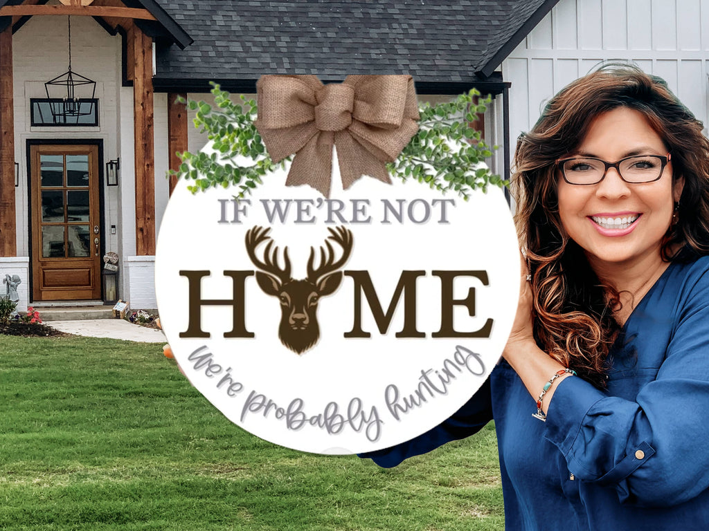A smiling woman stands in front of a house, holding a round sign with a deer head that reads, If were not home, were probably hunting. The sign is decorated with greenery, a bow, and cursive text.