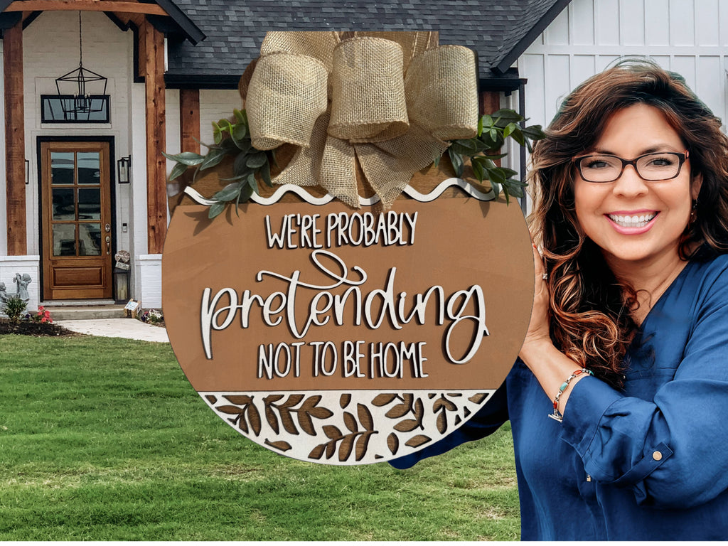A smiling woman stands outside a house beside the Were Probably Pretending Not To Be Home Door Sign, Door Wreath, decorated with greenery and burlap ribbon—a playful take on the standard door wreath.