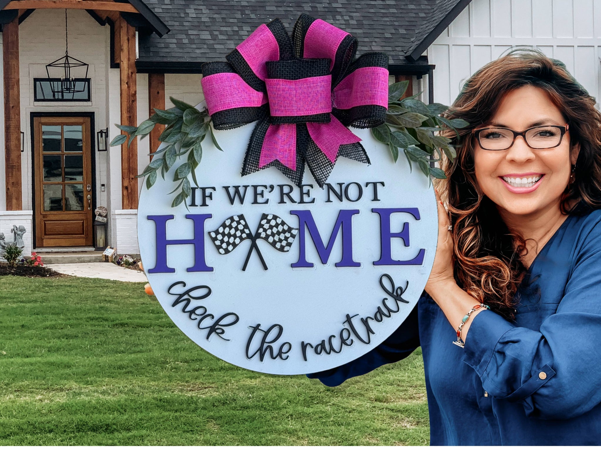 A woman with long brown hair and glasses smiles while holding a round sign with a large purple and black bow. The sign reads, If were not home, check the racetrack. A house is in the background.