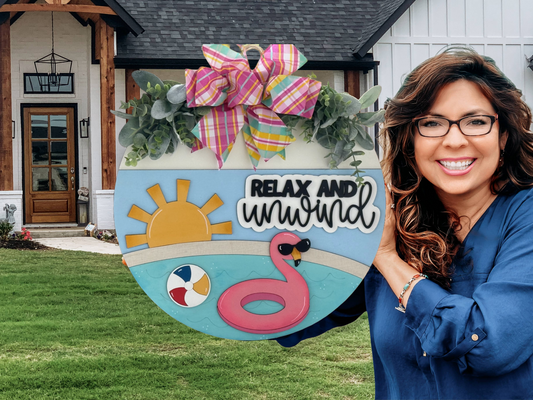 A smiling woman with long brown hair and glasses holds a round summer-themed sign that says Relax and Unwind, decorated with a sun, flamingo, beach ball, and a large colorful bow, standing in front of a house.