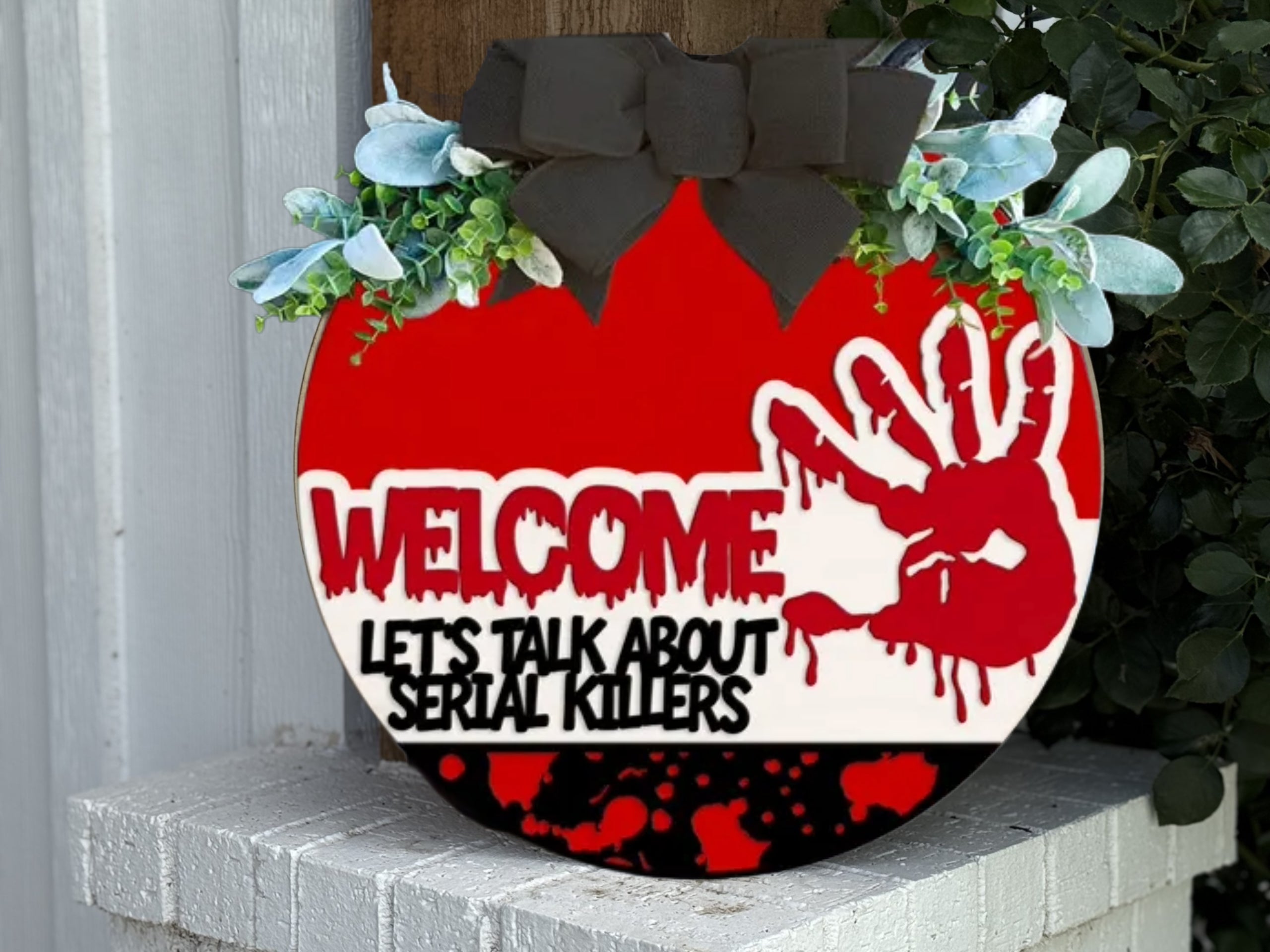 A round sign decorated with greenery and a black bow reads WELCOME LETS TALK ABOUT SERIAL KILLERS in bold letters. The design features a bloody handprint and splatters on a red and white background.