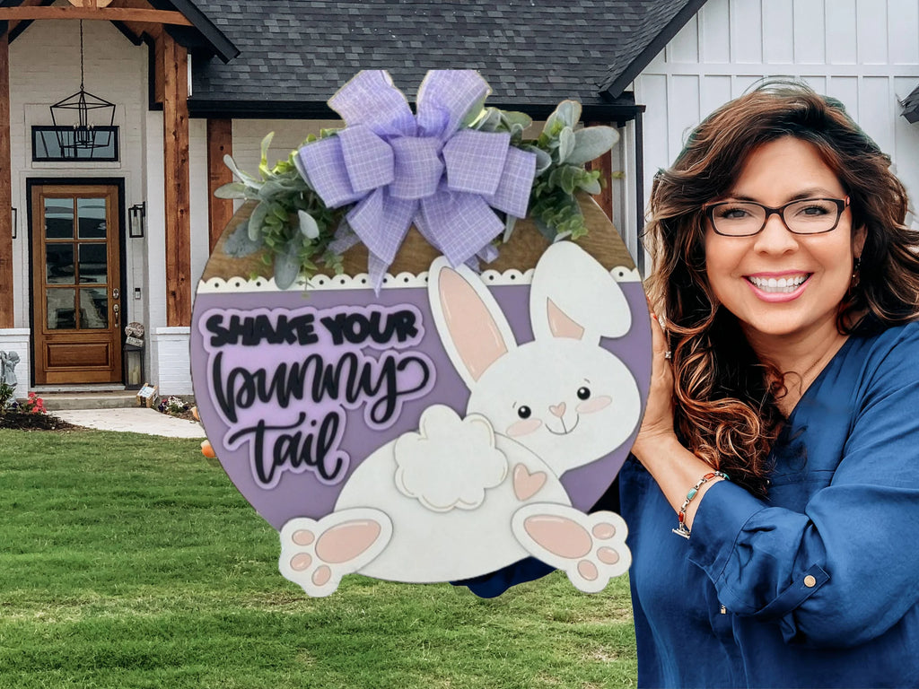 A smiling woman with wavy brown hair and glasses holds a round Easter sign with a bunny, purple bow, flowers, and the words “Shake your bunny tail.” She stands outside a white house with wood and brick accents, green grass, and a porch in the background.