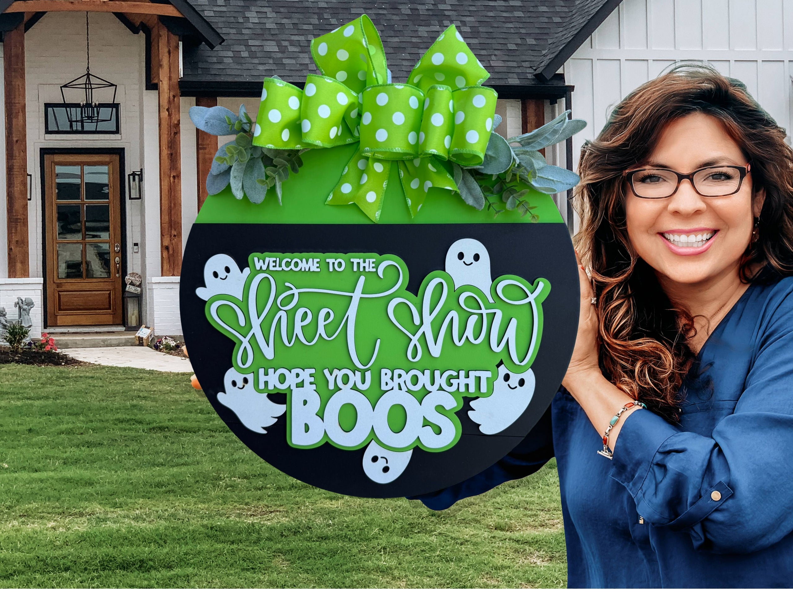 A round Halloween sign with a green top, polka-dot bow, leaves, and white ghosts, reading Welcome to the Sheet Show, hope you brought boos, displayed on a brick surface.