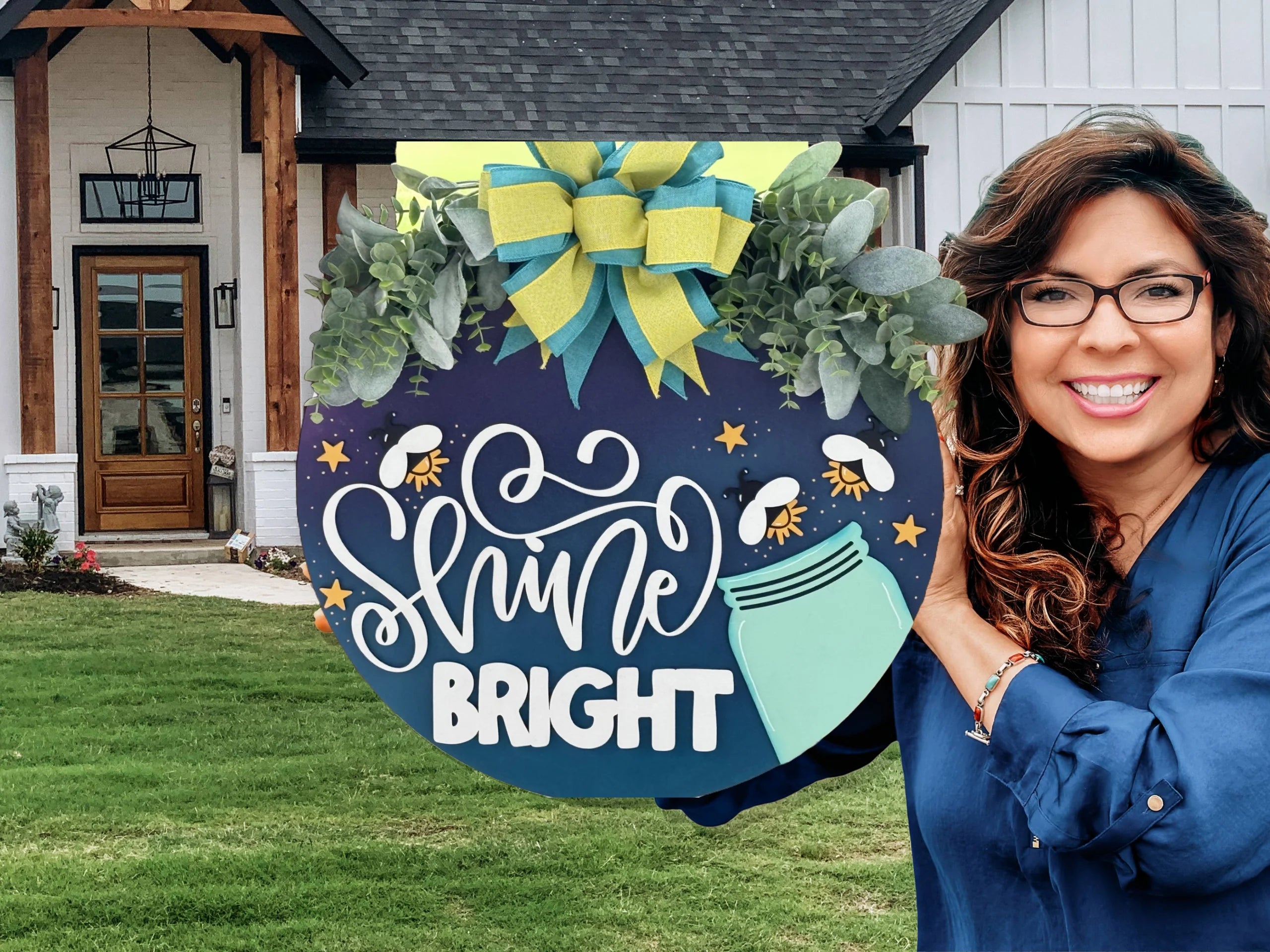 A smiling woman with wavy brown hair and glasses holds a large decorative sign reading “Shine Bright” with a mason jar, fireflies, blue–yellow bow, and green foliage, standing outside in front of a modern white house with a wooden porch.
