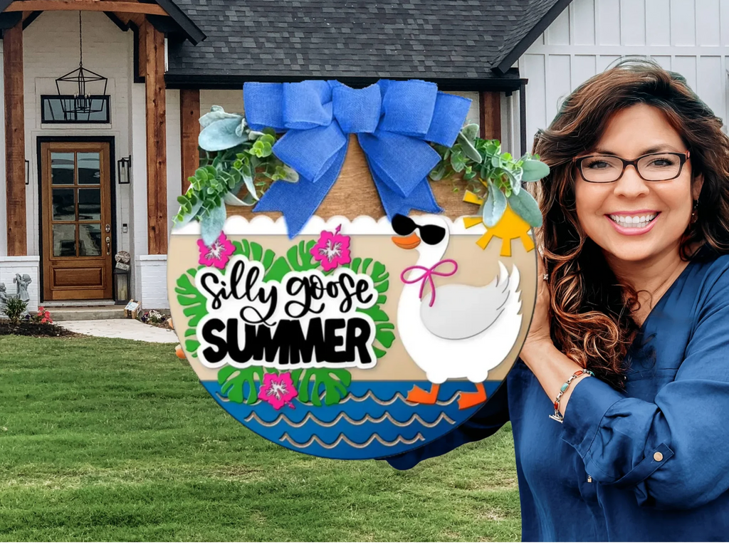 A smiling woman holding a colorful round sign that reads Silly Goose Summer with a goose wearing sunglasses, flowers, and a big blue bow, standing in front of a modern house with a green lawn.