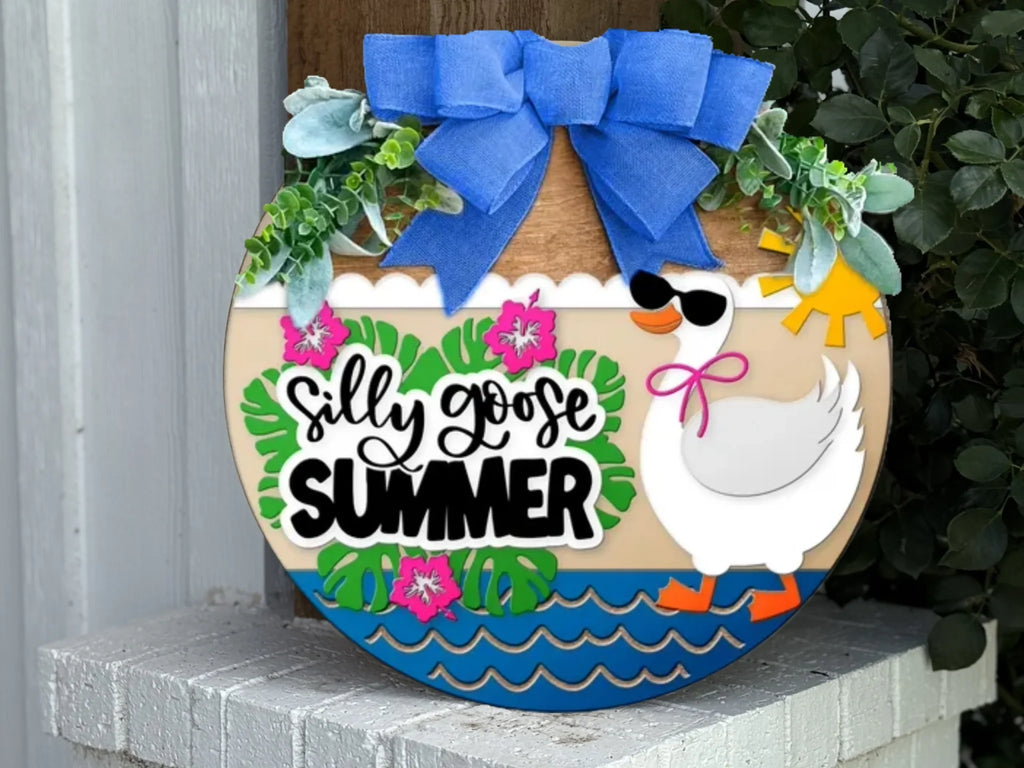 A round wooden sign decorated with leaves, flowers, and a duck wearing sunglasses and a pink bow, with the text Silly Goose Summer and a large blue ribbon at the top. The sign is displayed on a white brick surface.